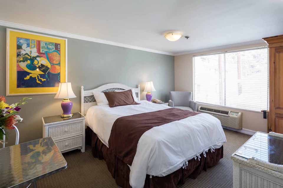 Hotel room with a white bed, brown accent, two wicker nightstands, purple lamps, and a colorful abstract painting by the bed.